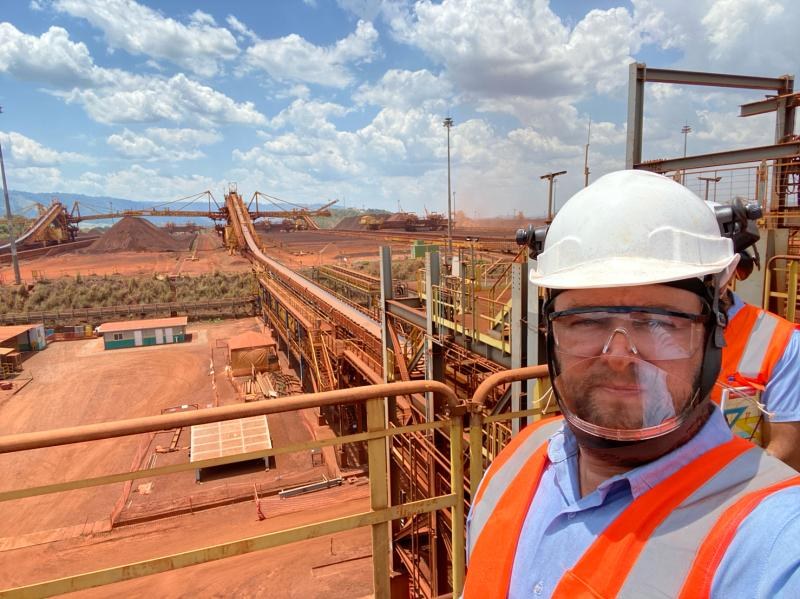 Gustavo de capacete, óculos e colete refletivo em passarela de planta de minério de ferro, com pilhas, correias e empilhadeira-recuperadora ao fundo